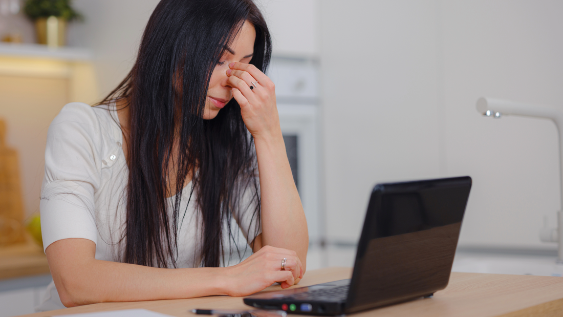 A white woman with black hair wearing a white blouse sits at a laptop computer at her office desk. She's pinching her nose between her fingers. 