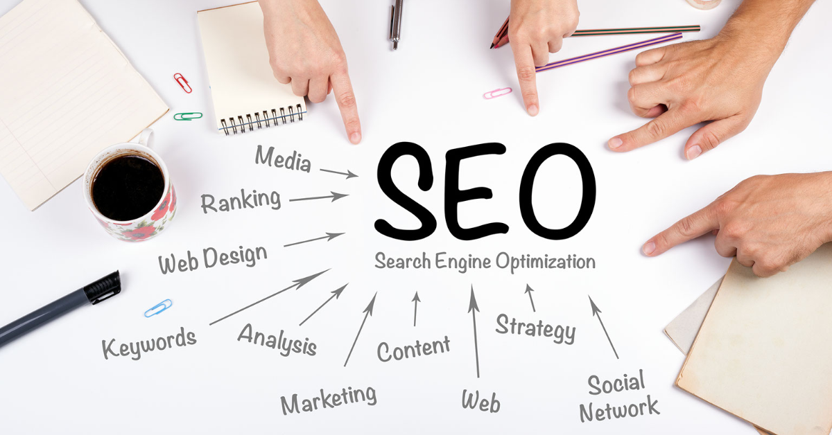 Several hands point to the words SEO on a table in front of them, with other words around it to describe what Search Engine Optimization does.