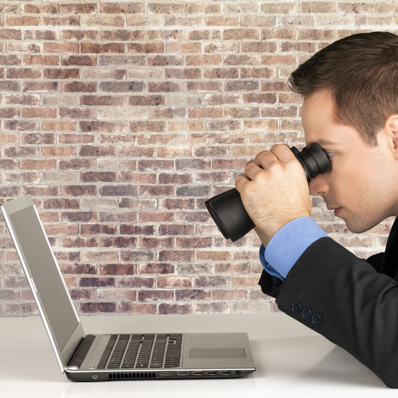 Man looking through a pair of binoculars at a laptop. 