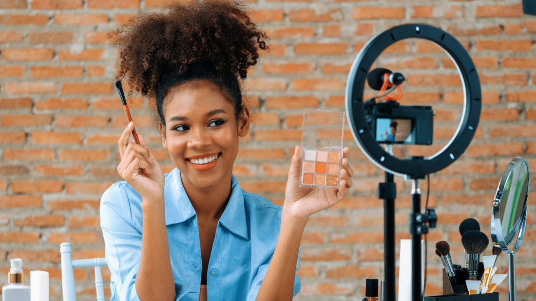 A Black woman wearing a blue shirt holds a makeup pallet and brush and demonstrates how to use it to her social media followers. 