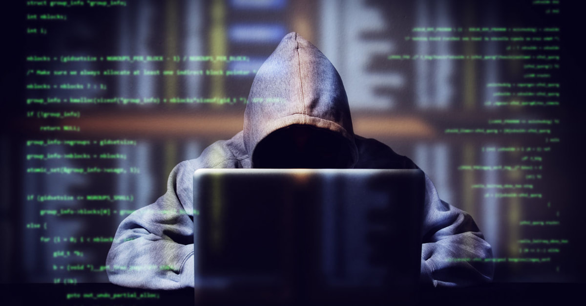 A hacker wearing a hoodie sits in front of a laptop. Computer code appears behind him.