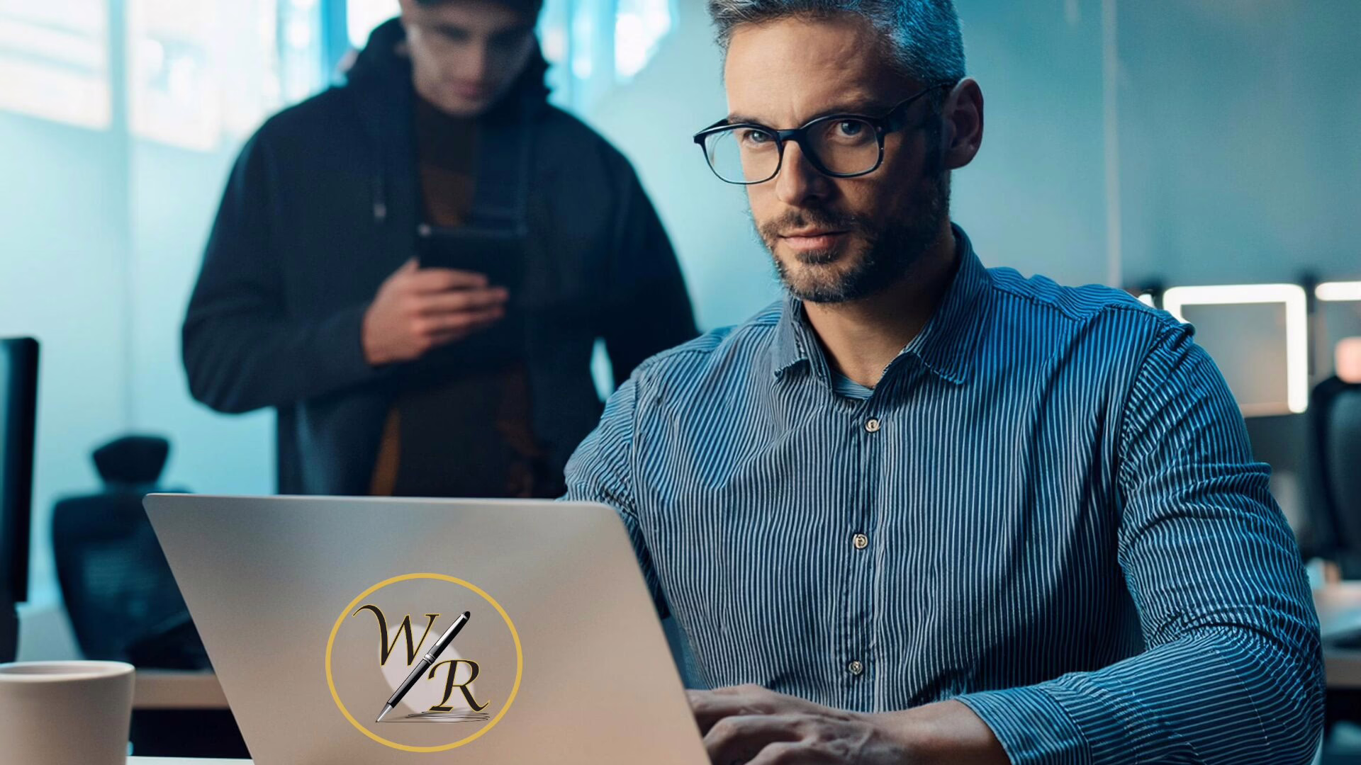 A man with glasses sits at a computer using LinkedIn, while a scammer lingers around behind him.