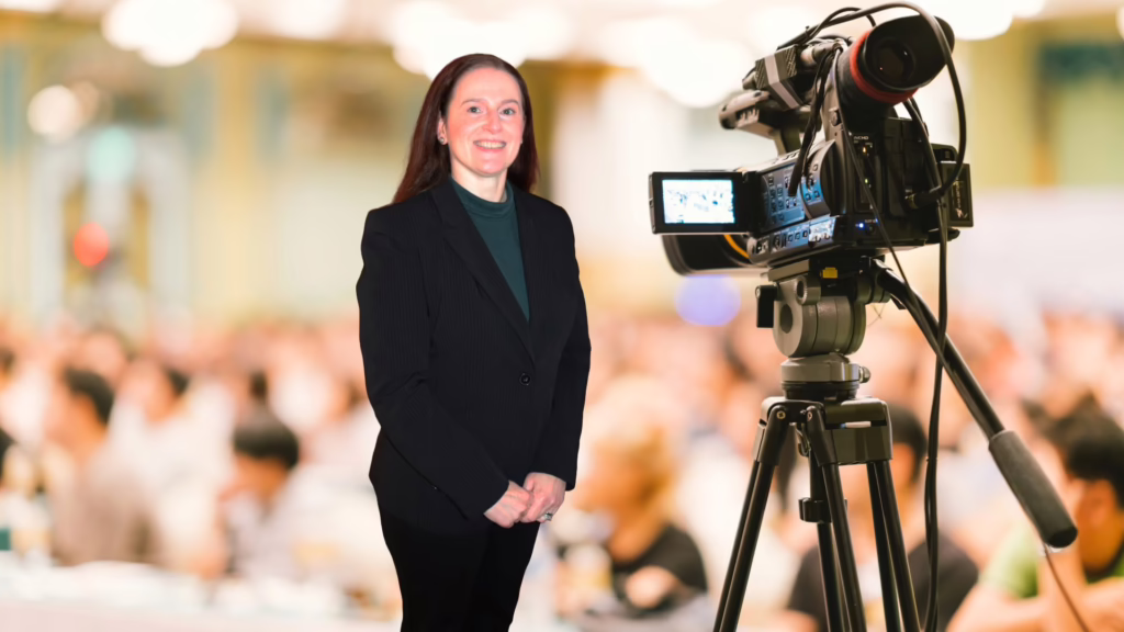 Shari Berg of The Write Reflection stands in front of a camera with a public meeting happening in the background.