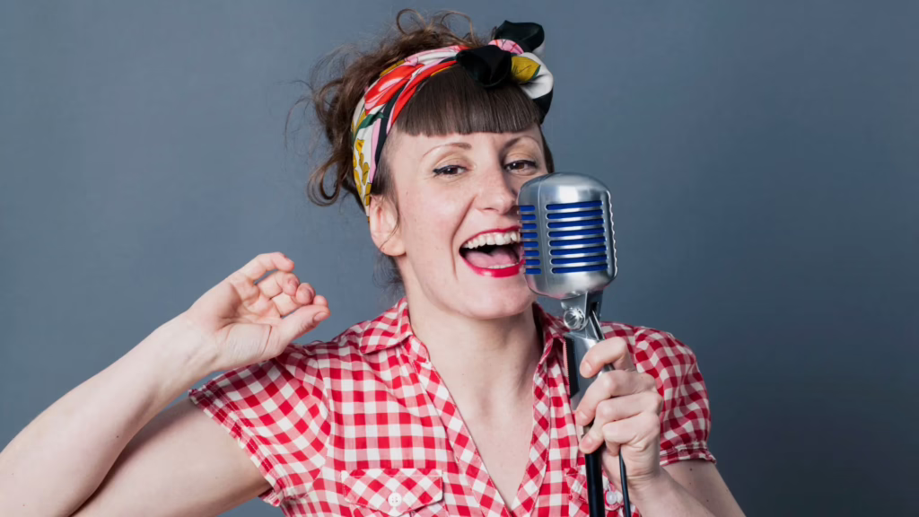 A red-headed woman wearing a bright red shirt and lipstick grabs a microphone and speaks into it. 
