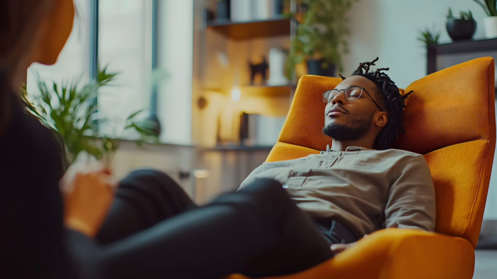 A black man relaxes in an orange chair as he receives hypnosis-based coaching. 