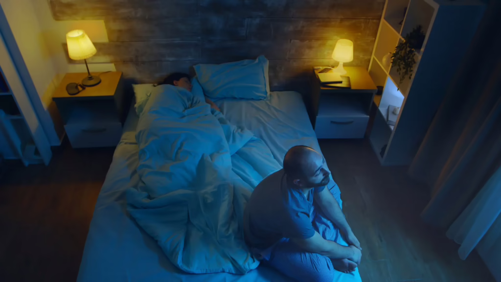 A man sits on the edge of his bed, struggling with insomnia as his wife sleeps soundly. 