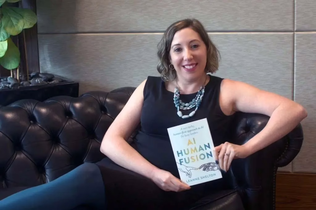 Author Leanne Shelton sits on a couch smiling while holding her new book, "AI Human Fusion."